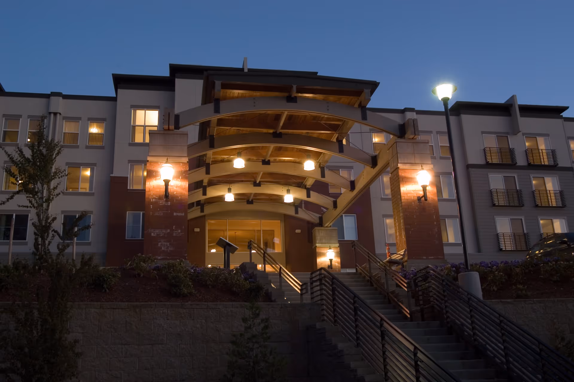 Exterior view of The Heights at Columbia Knoll building entrance at dusk, featuring a covered walkway with wooden beams, illuminated lamps, stairs leading up to the entrance, and multiple windows with lights on inside.