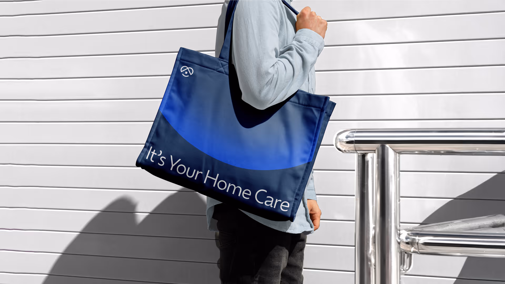 A person outdoors carries a blue bag that reads It's Your Home Care.