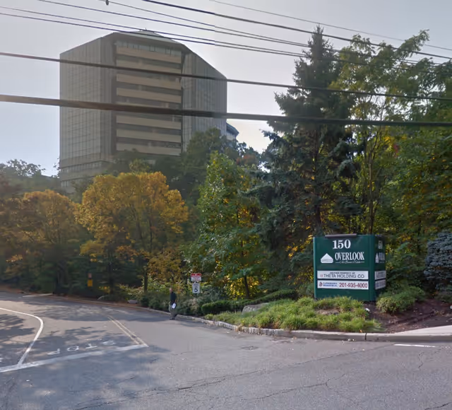 Tall modern building behind trees with a green sign beside a curved road.