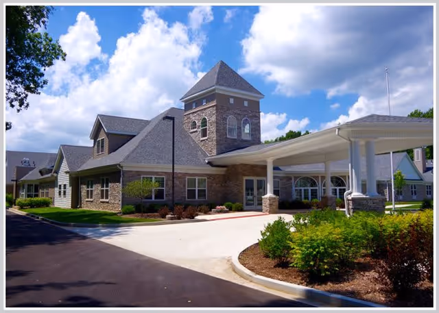 Front exterior of a stone-faced senior health facility with a covered porte-cochère, landscaped driveway, and tower under a blue sky.