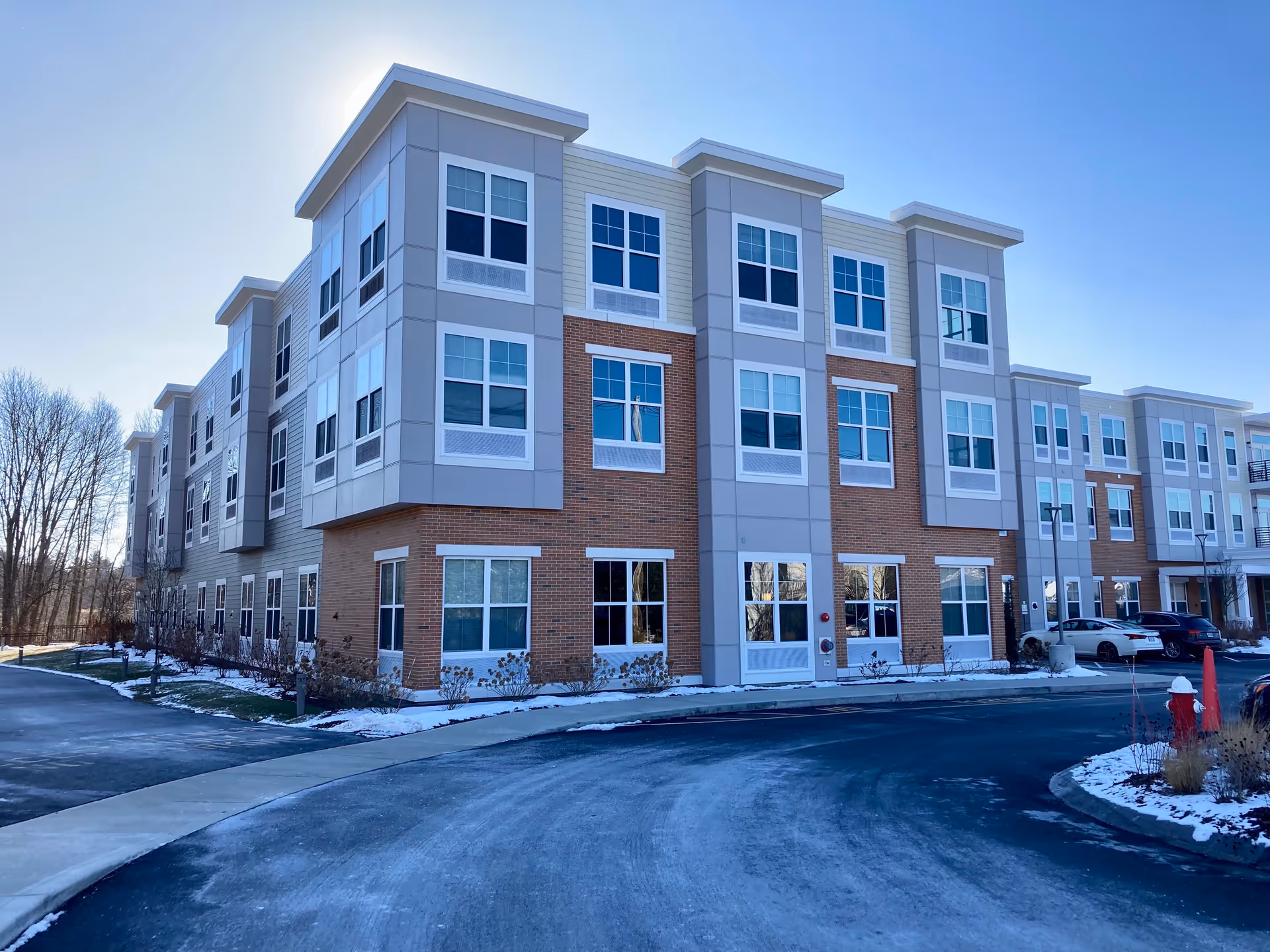 Three-story modern senior living building with brick and gray paneling, many windows, a curved driveway and parked cars in front.