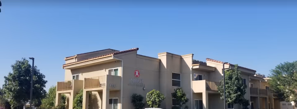 Two-story beige senior living building with balconies and landscaping under a clear blue sky.