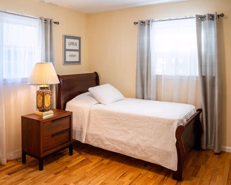 A furnished bedroom in a senior living facility with a wooden bed, nightstand, and lamp.