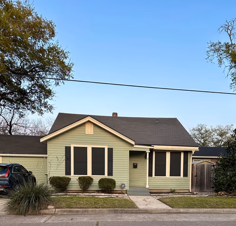 Front view of a pale green, single-story house with shutters and shrubs.