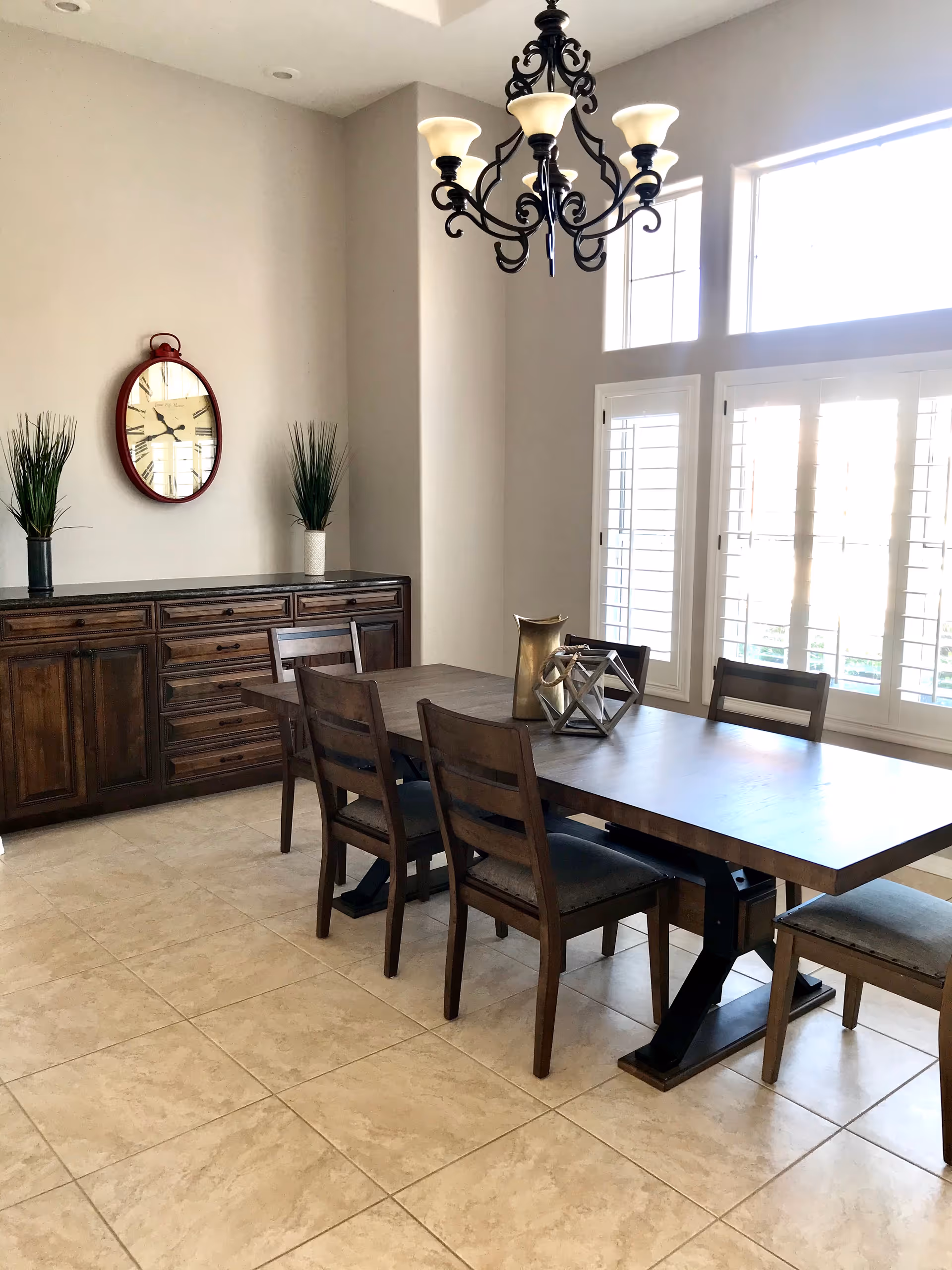 A bright dining room with a large wooden dining table surrounded by six chairs. The room features a dark wooden sideboard with two tall green plants in vases and a large round clock hanging above it. There are large windows with white shutters allowing natural light to fill the space, and a decorative chandelier hangs from the ceiling.