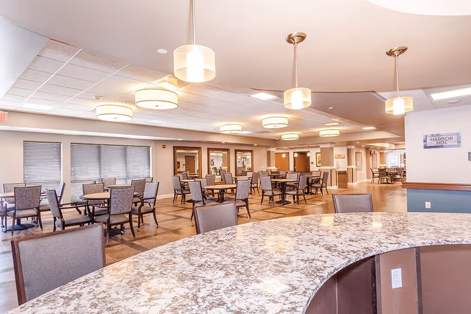 A spacious dining area in an assisted living facility with multiple tables and chairs arranged neatly. The room features large windows with blinds, modern pendant and ceiling lights, and a curved granite countertop in the foreground.