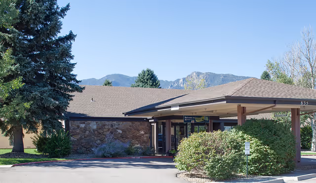 Front exterior view of Mountain View Post Acute facility with a covered entrance, surrounded by greenery and trees, with mountains visible in the background under a clear blue sky.