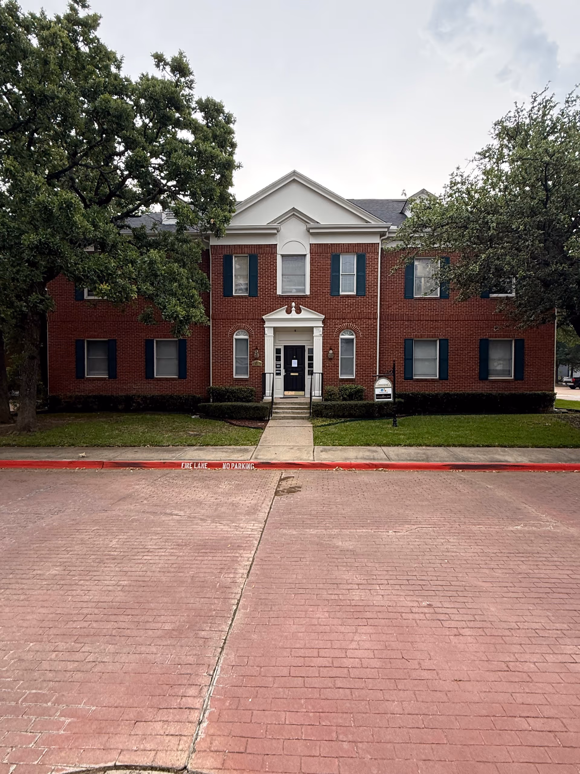 Brick two-story building with a central entrance, surrounding trees, and a sidewalk leading to stairs.