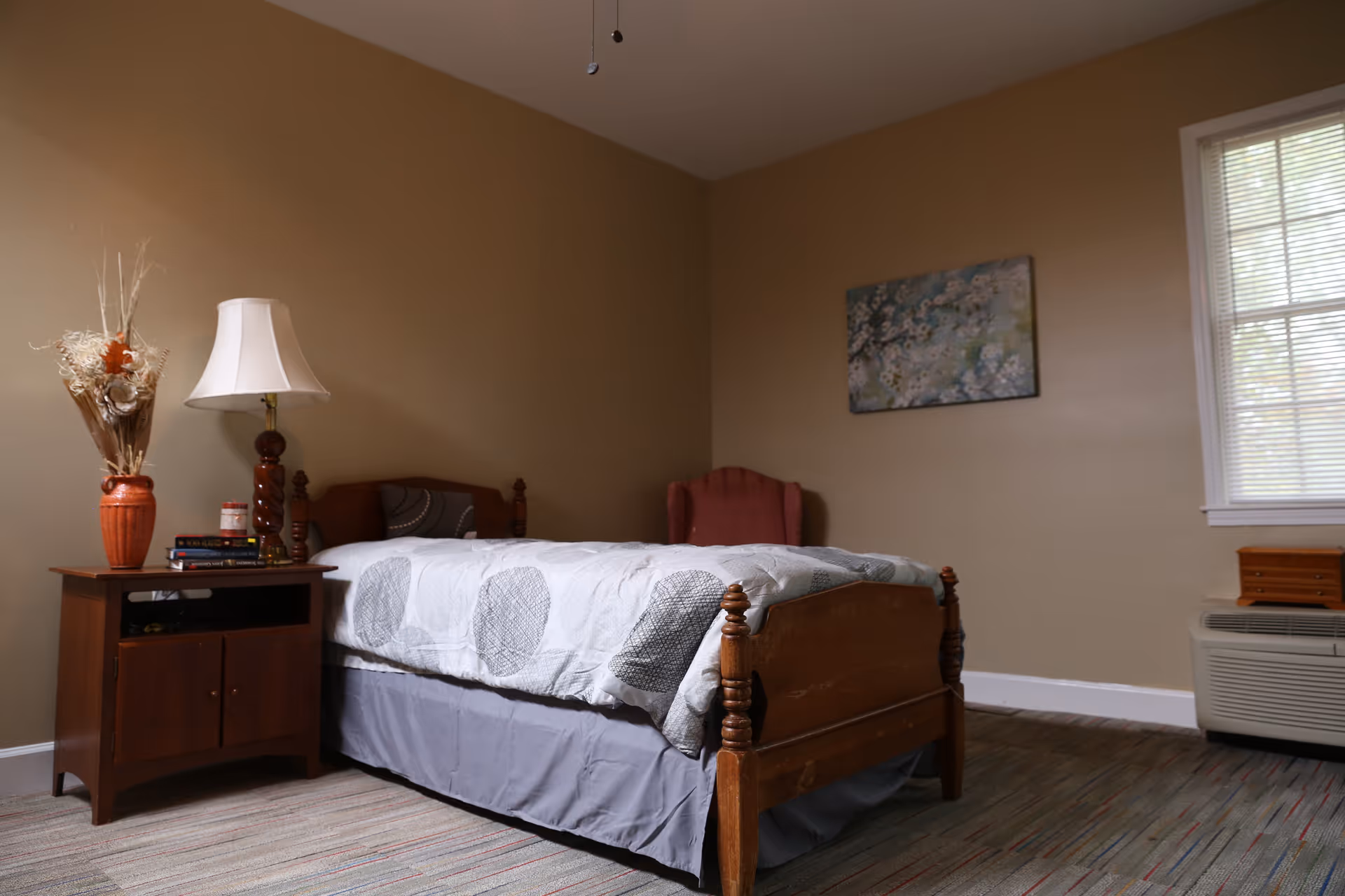 A simple, well-lit bedroom featuring a wooden bed, nightstand, lamp, and window.