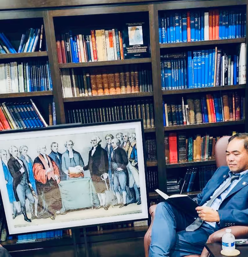 Man in a blue suit sits reading a book beside a large bookshelf in a library.