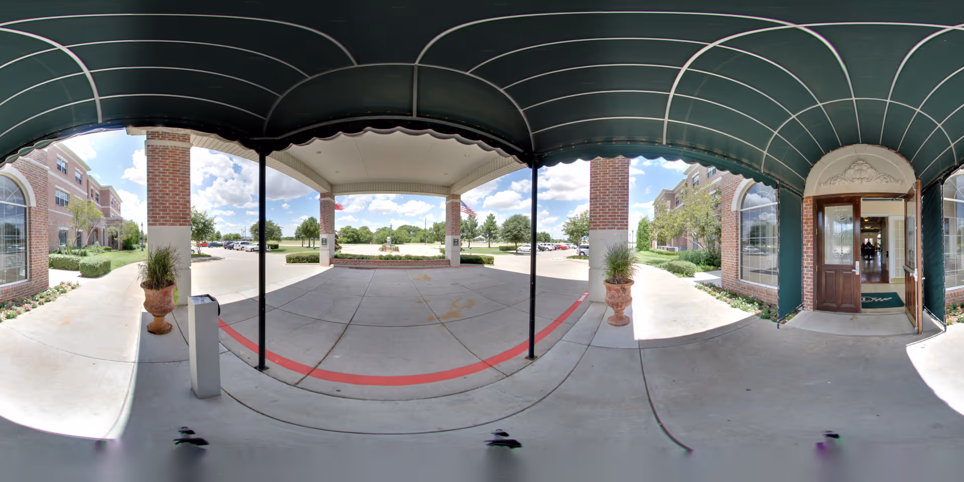 Covered entrance area of a senior living facility with a green awning overhead, brick pillars, potted plants, and a view of the parking lot and landscaped grounds. The entrance door to the building is open, showing a glimpse of the interior.