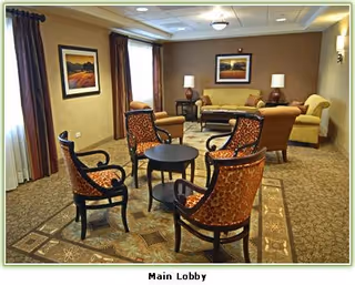 A cozy senior living community lobby with a seating area featuring four patterned armchairs around a small round table on a decorative rug. In the background, there are two sofas, two lamps on side tables, framed artwork on the walls, and large windows with curtains allowing natural light.