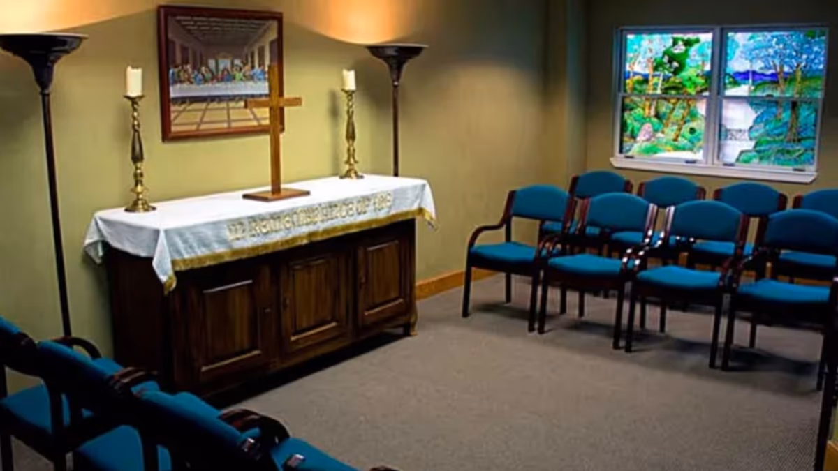A small chapel room with rows of blue cushioned chairs arranged facing a wooden altar. The altar is covered with a white cloth and has a wooden cross and two candles on it. On the wall above the altar is a framed picture, and to the right, there is a window with colorful stained glass depicting a nature scene.