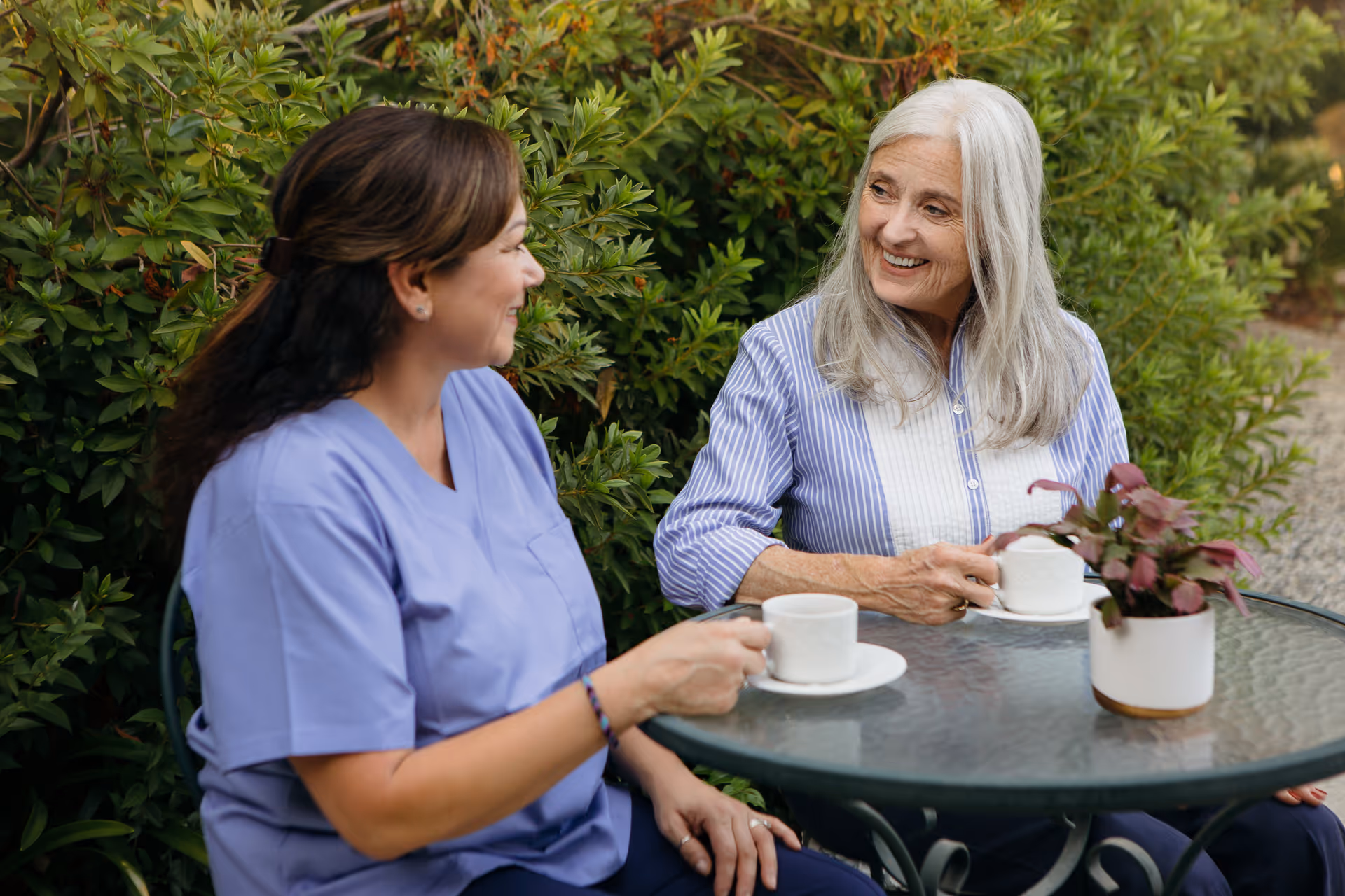 Two women chat at an outdoor table, caregiver in scrubs and an elderly resident.