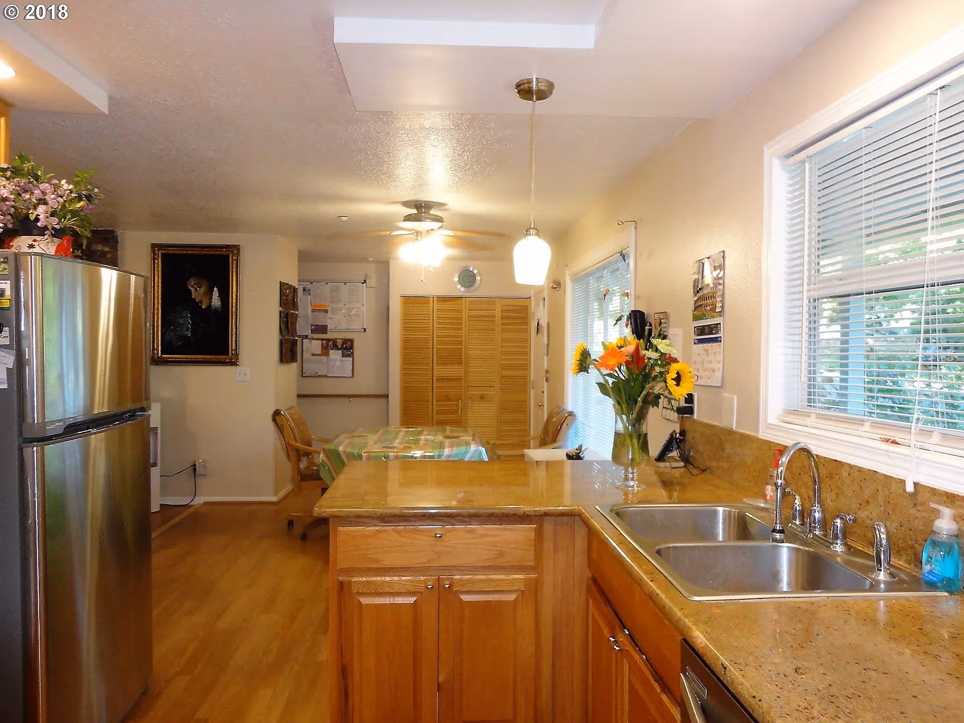 Bright open kitchen with wooden cabinets, a stainless refrigerator, double sink, and a dining table in the background.