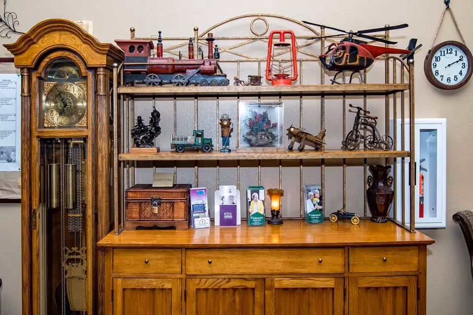 A wooden cabinet with shelves displaying various decorative items including a model train, a red lantern, a model helicopter, small figurines, a wooden chest, brochures, and a small lamp. To the left of the cabinet is a tall wooden grandfather clock. A round wall clock hangs on the right side above a fire extinguisher cabinet.