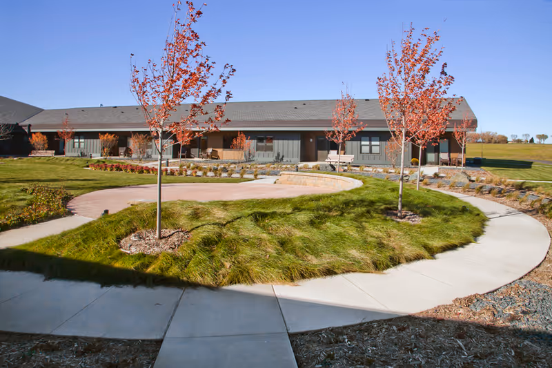 Single-story senior living building exterior with curved walkway, landscaped circular lawn, and young trees.