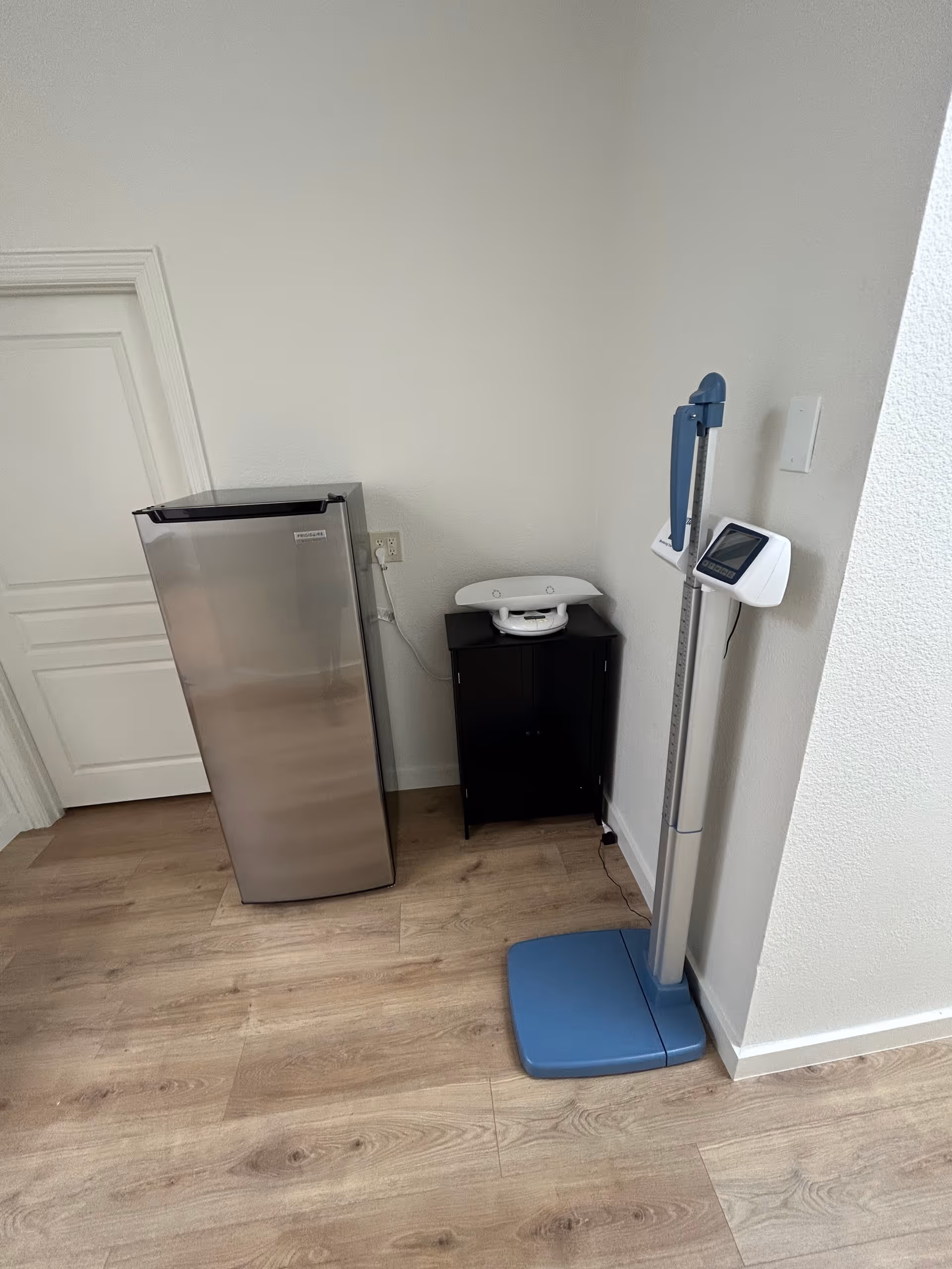 A small room corner with a stainless fridge, a digital scale, and a cabinet.