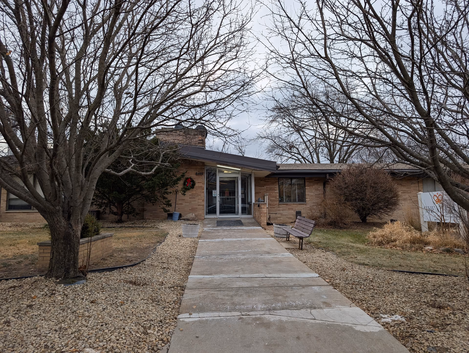Walkway leading to the front entrance of a low, single-story brick building with leafless trees, a bench, and a wreath on the door.