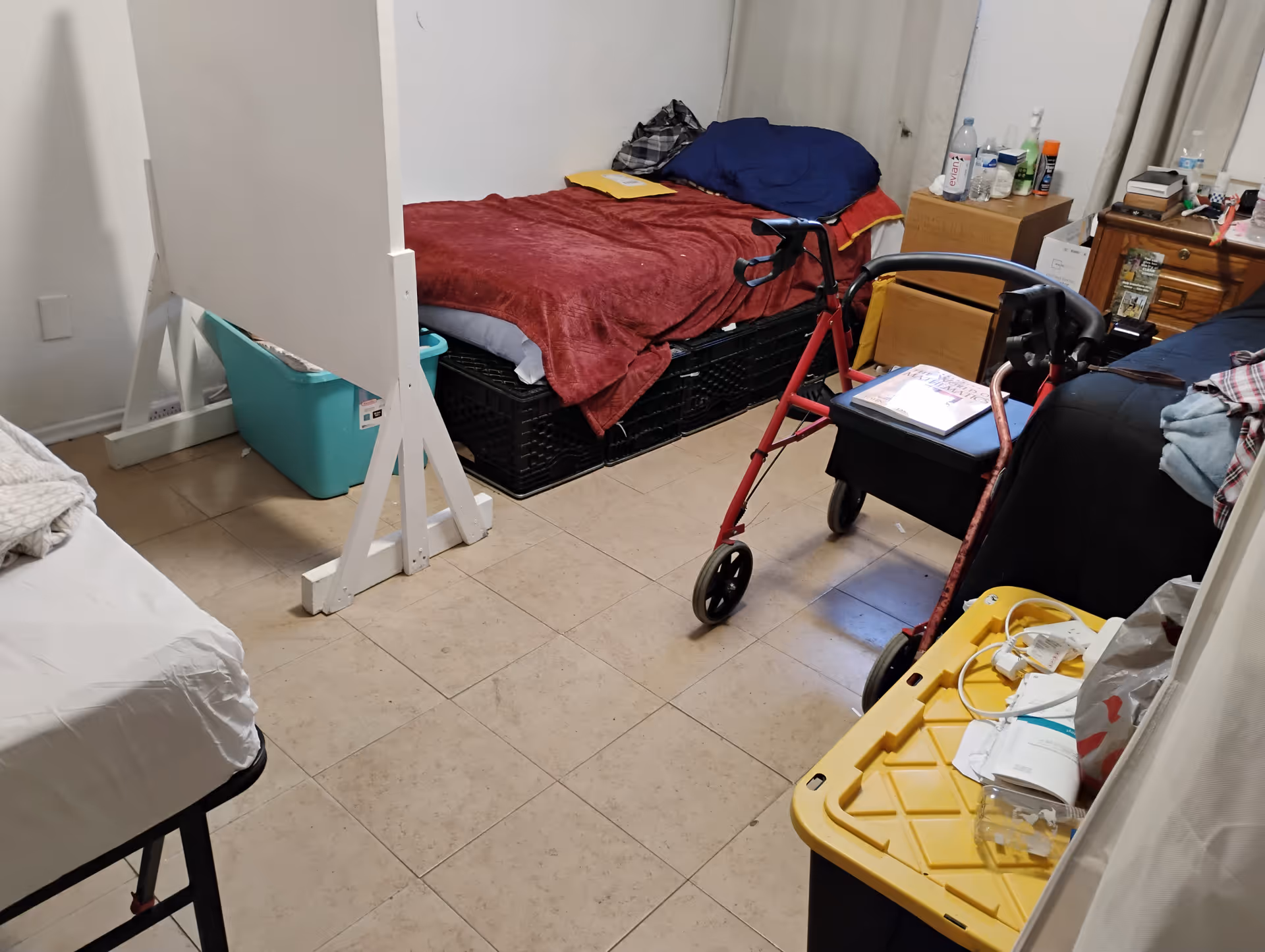 Small, cluttered resident room with beds, a walker, and medical supplies.