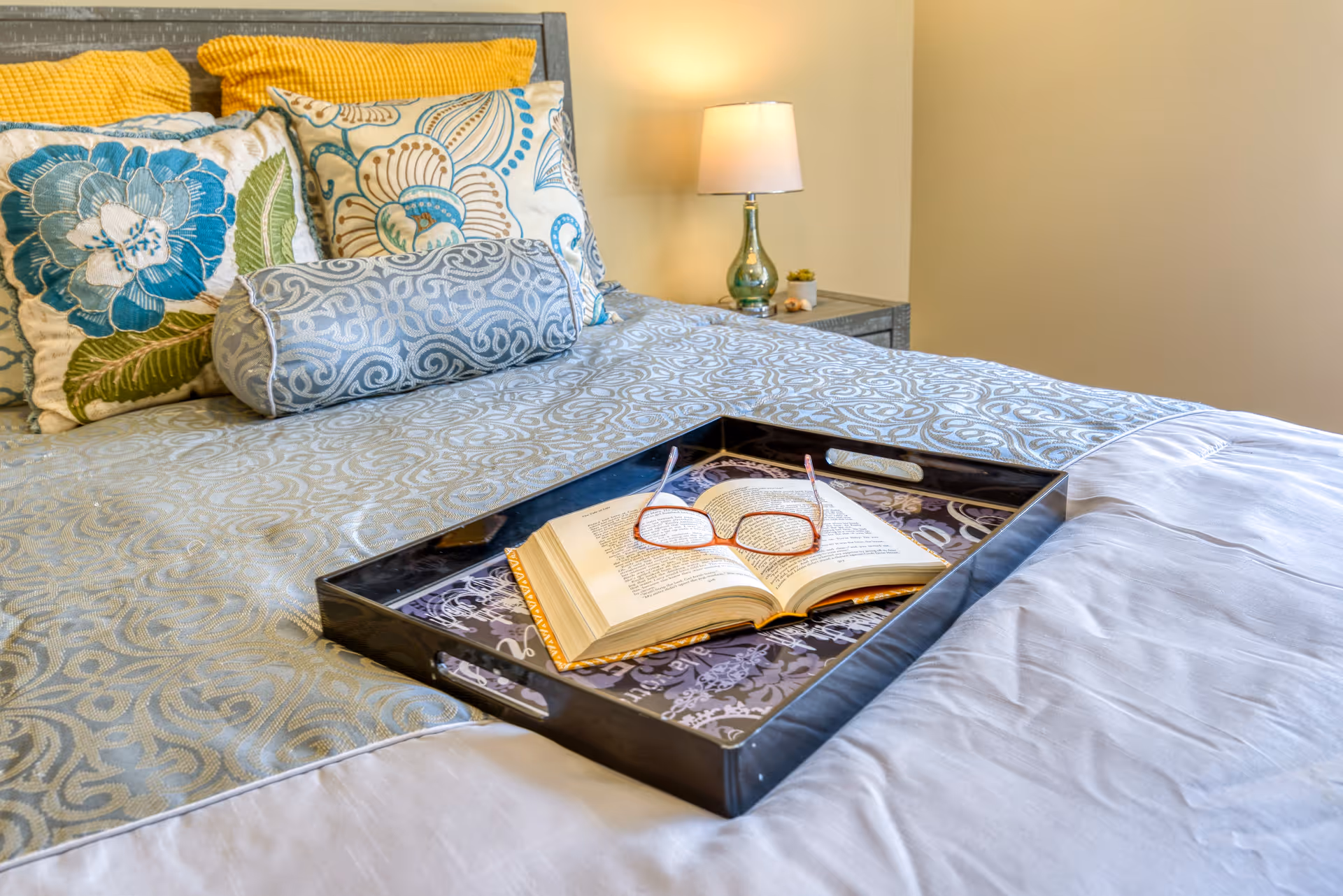 A neatly made bed with decorative pillows in blue, green, and yellow patterns. On the bed is a black tray holding an open book and a pair of reading glasses. A bedside table with a lamp and small plant is visible in the background.