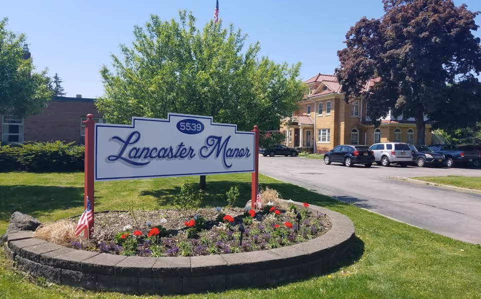 Front exterior view of Lancaster Manor sign with a flower bed and campus building in background.