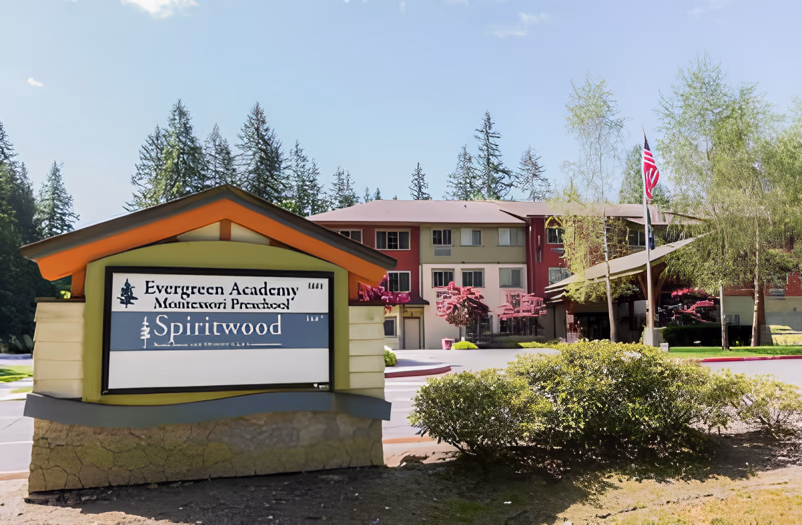 A sign reading 'Spiritwood' and 'Evergreen Academy Montessori Preschool' stands in front of a multi-story residential building with landscaping and an American flag.