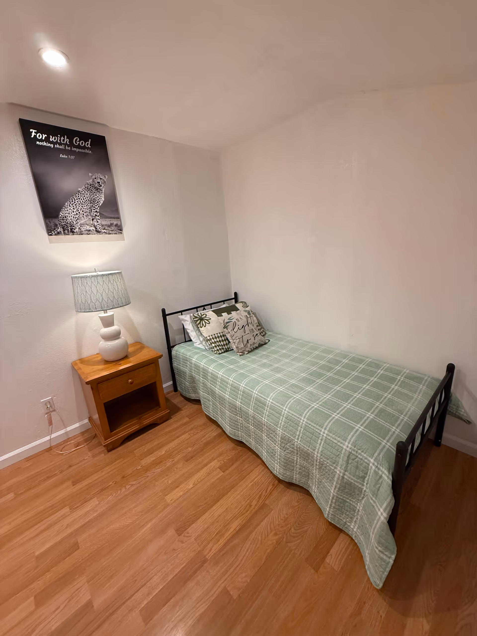 Single bed with green plaid cover, nightstand, lamp, and framed wall art in a small bedroom.