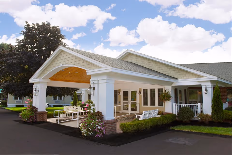 Exterior view of the entrance to Edgewood Center, featuring a covered drop-off area with white columns, benches, hanging flower baskets, and well-maintained landscaping under a partly cloudy sky.