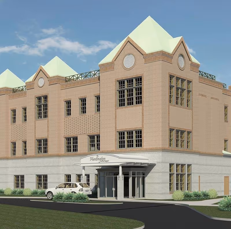 Exterior view of a three-story assisted living facility building with beige and light brown brick walls, multiple windows, and a white entrance canopy labeled 'Huntington Assisted Living'. A white car is parked near the entrance, and there is a paved driveway and green landscaping around the building under a blue sky.
