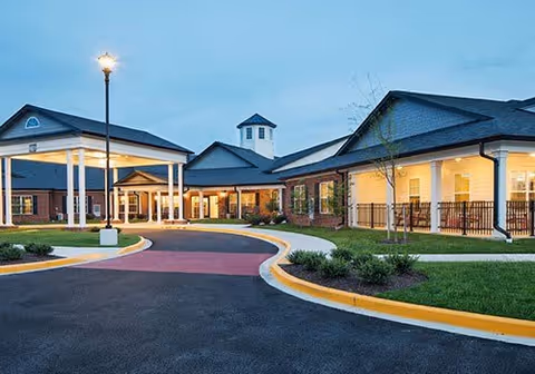 Front entrance of a single-story brick senior living facility with a covered porte-cochere, circular driveway, lamp post, and landscaped grounds at dusk.