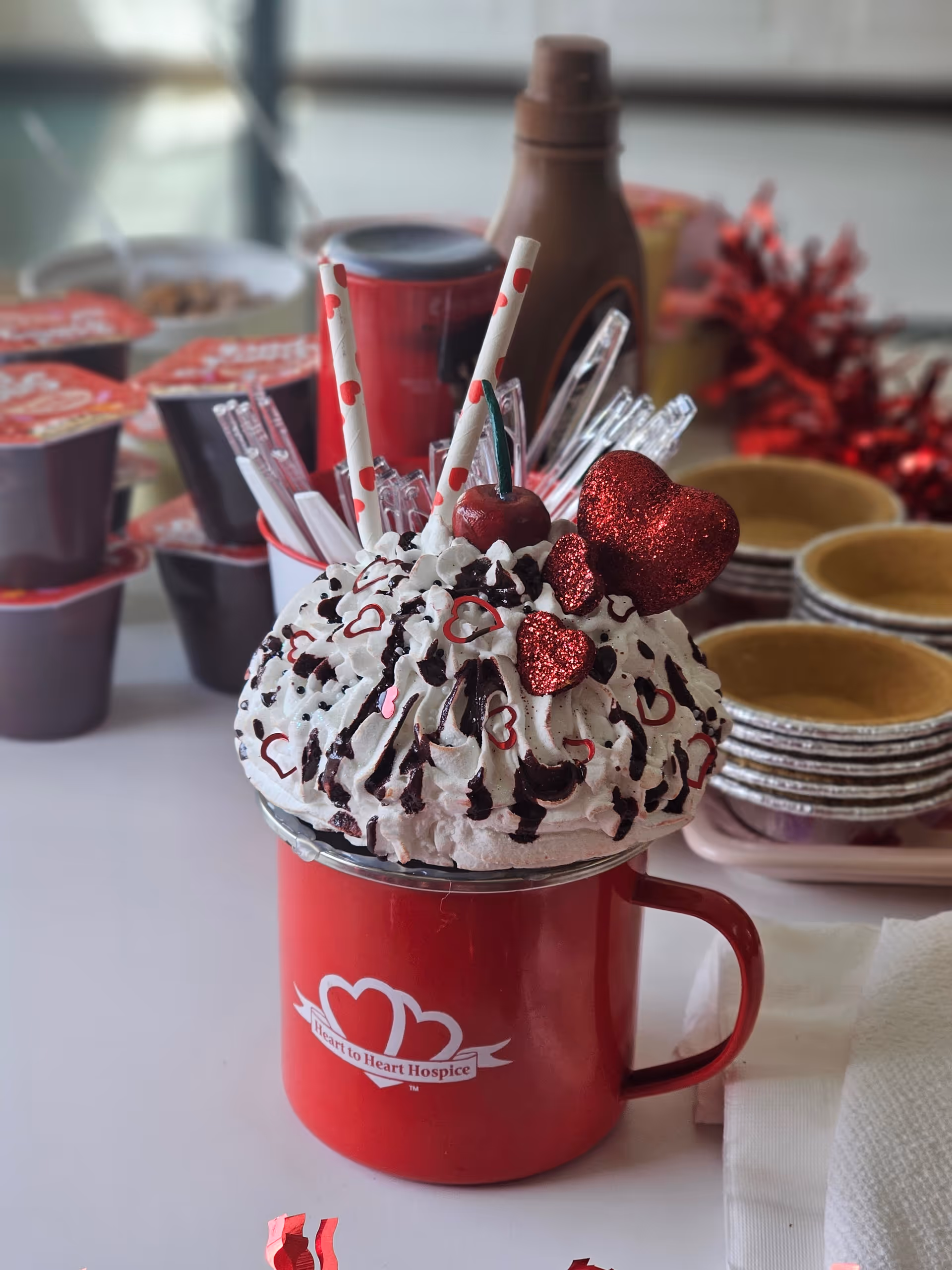 Red mug with whipped cream, chocolate drizzle, and red heart decorations on a dessert setup.