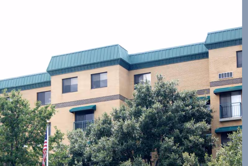 Exterior view of a multi-story brick building with green roof accents and several windows, partially obscured by leafy trees in the foreground.