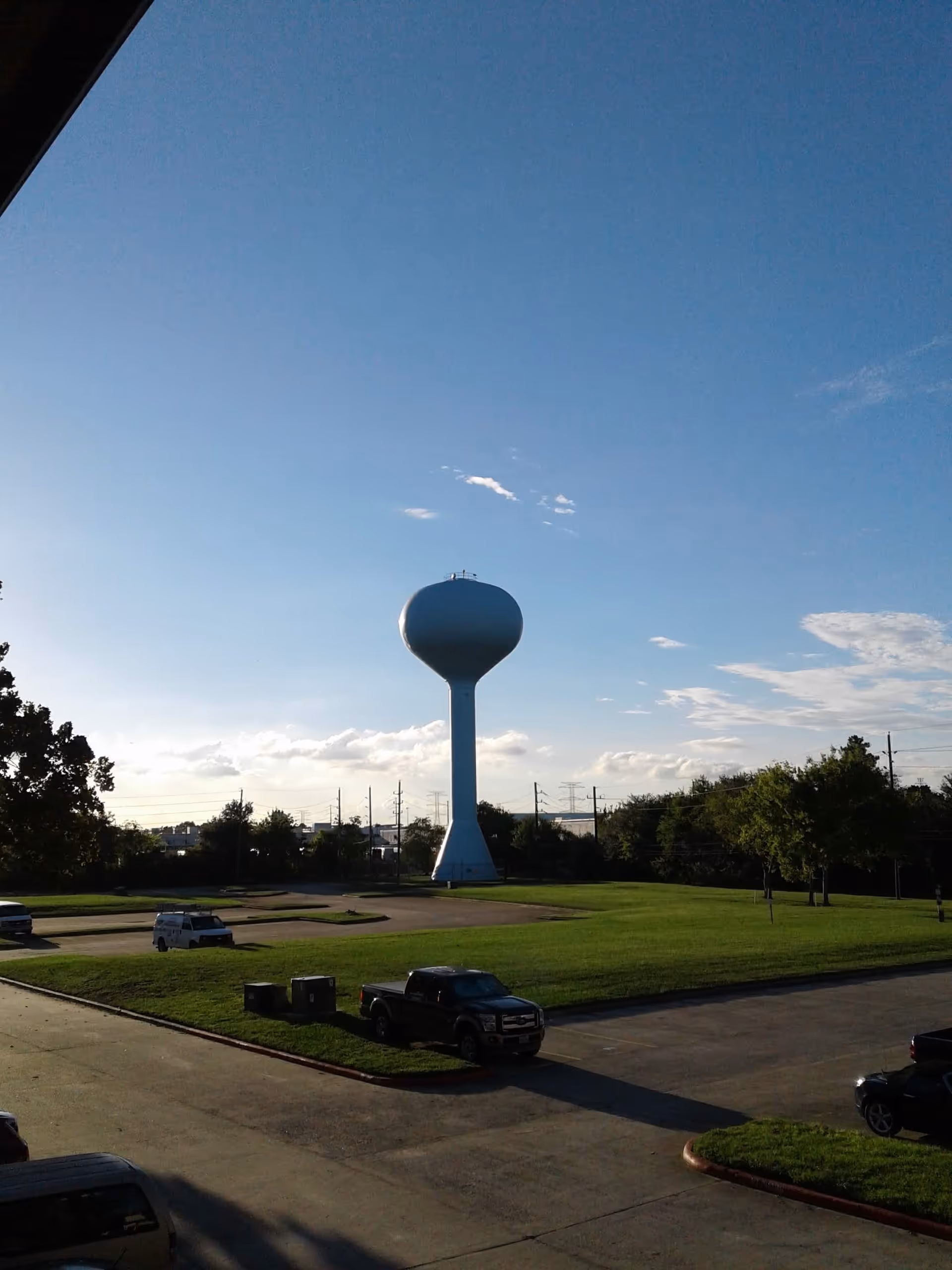 A large water tower rises beside a grassy lot with parked cars under a blue sky.