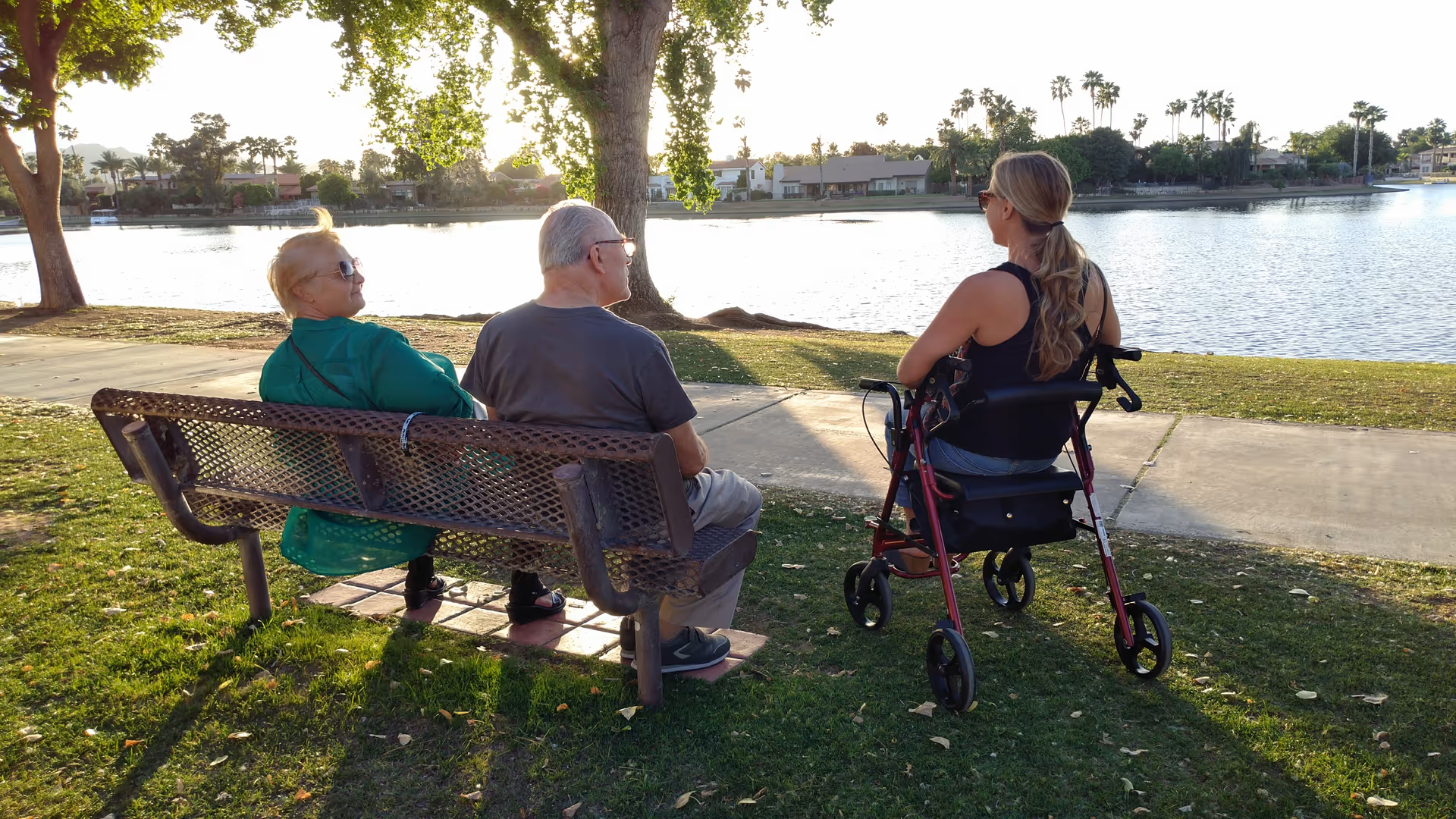 Two elderly people sitting on a park bench and a woman with a walker sitting beside them, all facing a lake with trees and houses in the background during sunset.