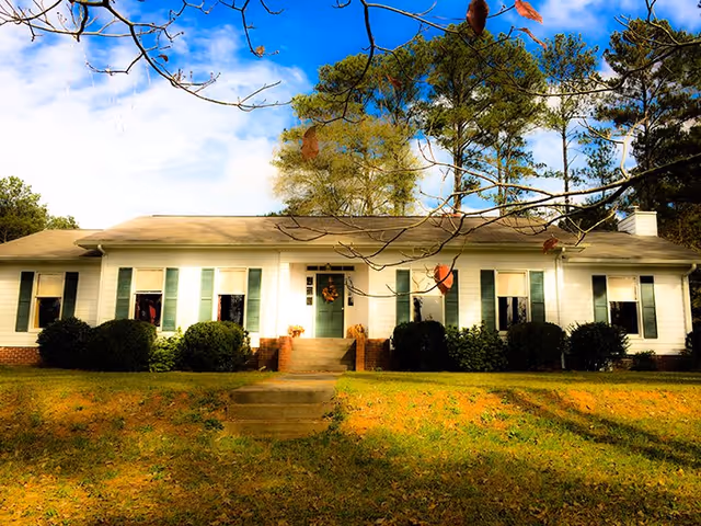 Single-story white building with green shutters and front steps, surrounded by trees.