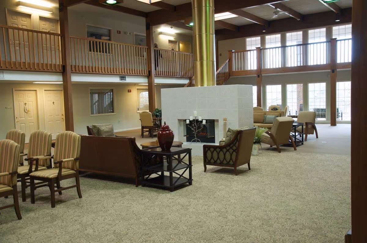 Spacious senior living community lounge area with multiple seating arrangements including armchairs and sofas around a central fireplace. The room features large windows allowing natural light, a high ceiling with exposed wooden beams, and a second-floor balcony overlooking the space.
