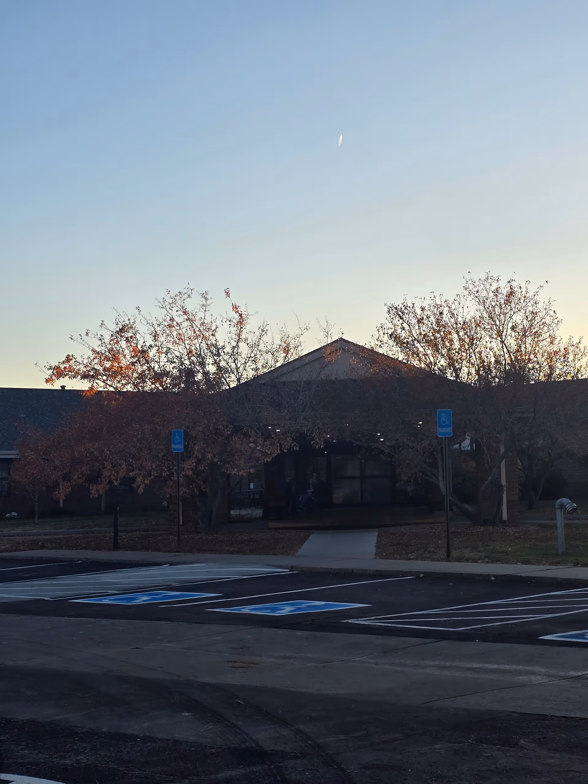 Exterior view of the Brookside Inn building at dusk with a clear sky. The entrance is partially obscured by trees with autumn leaves. In front of the building, there is a parking lot with several marked handicap parking spaces.