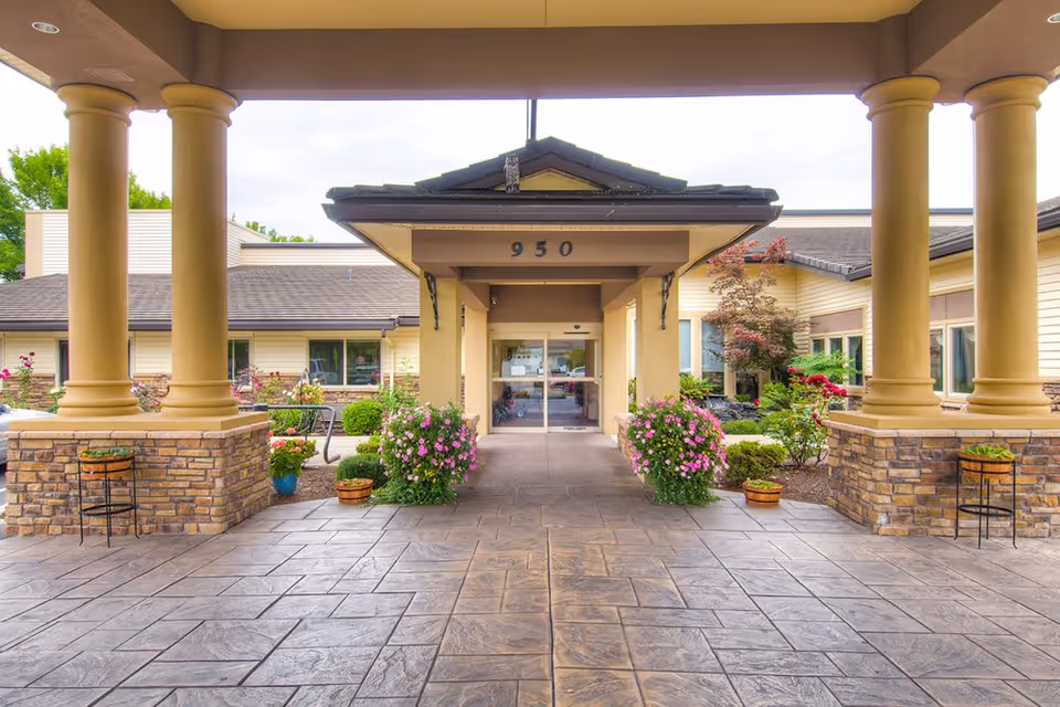 Covered entrance of a retirement community building with large columns, potted flowers, and glass double doors beneath the number 950.