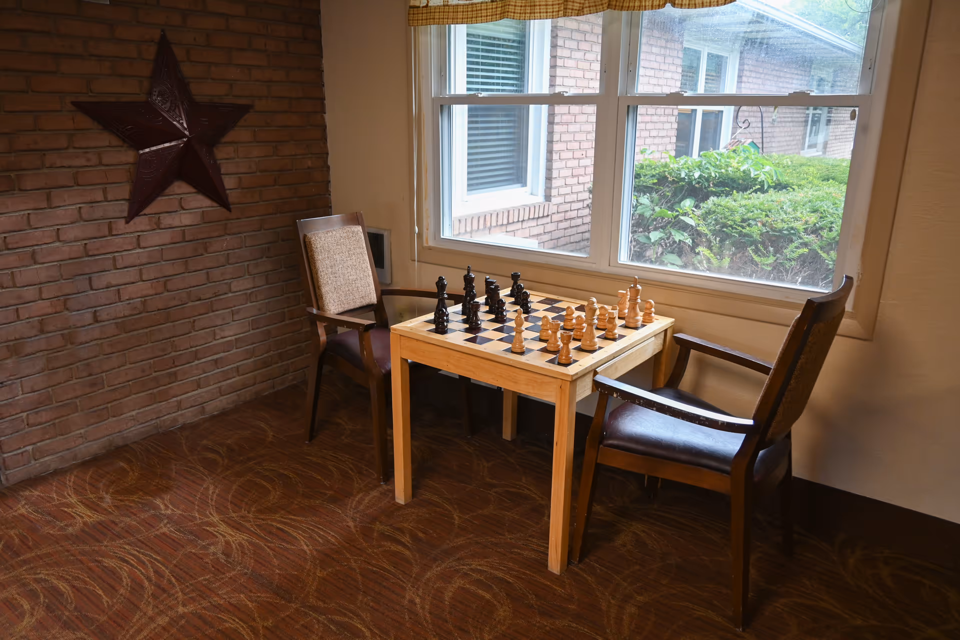 Two chairs sit around a chessboard on a wooden table by a window.