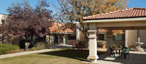 Sunny courtyard with a covered patio, table and chairs, a fountain, lawn and tiled-roof building surrounded by trees.