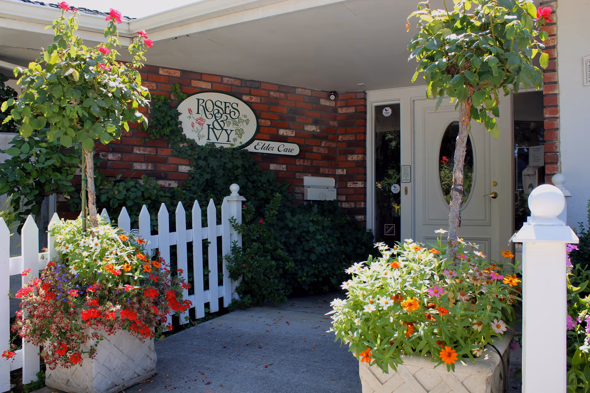 Front entrance of Roses & Ivy assisted living with a white picket fence, flowering planters, and a brick wall sign.