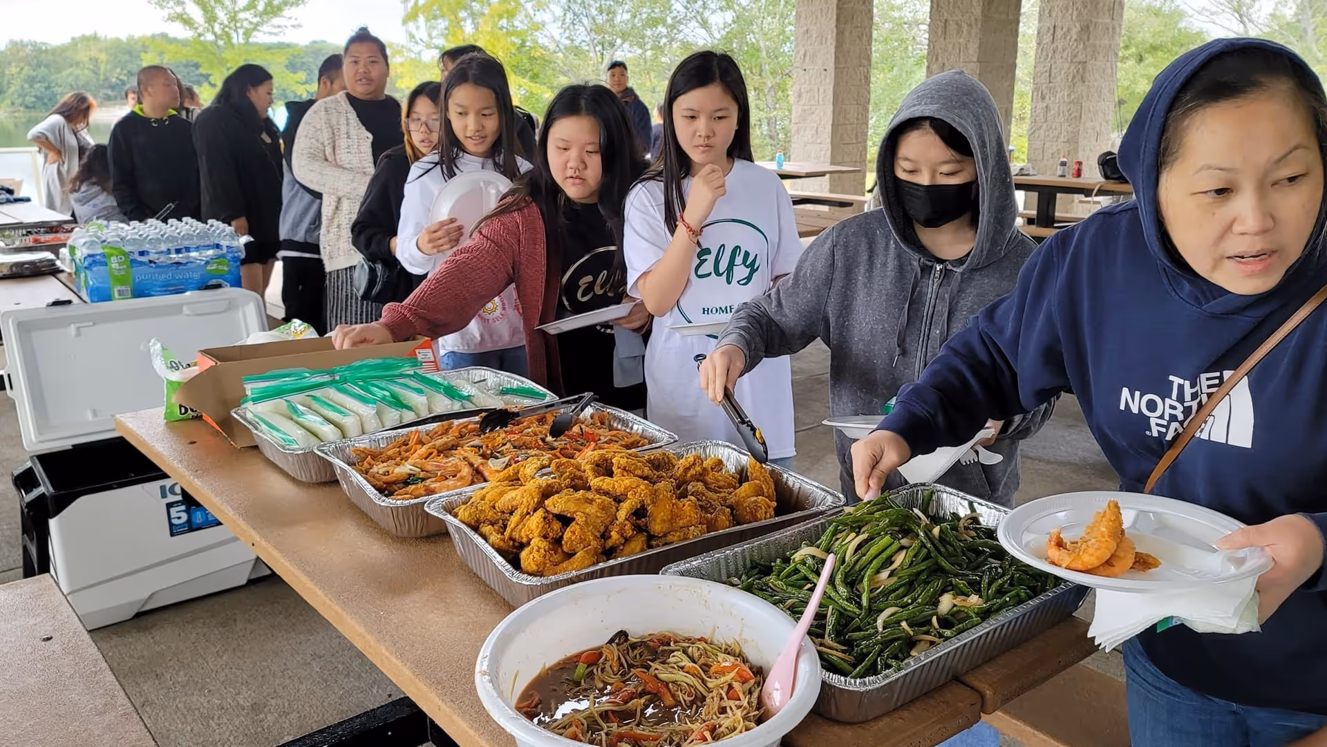 Group of people line up for food at an outdoor Elfy Home Care buffet.