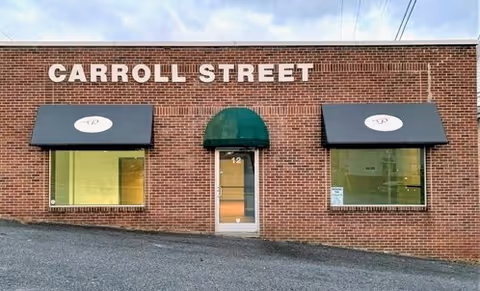Brick building exterior with Carroll Street sign, two windows with awnings, central glass door.