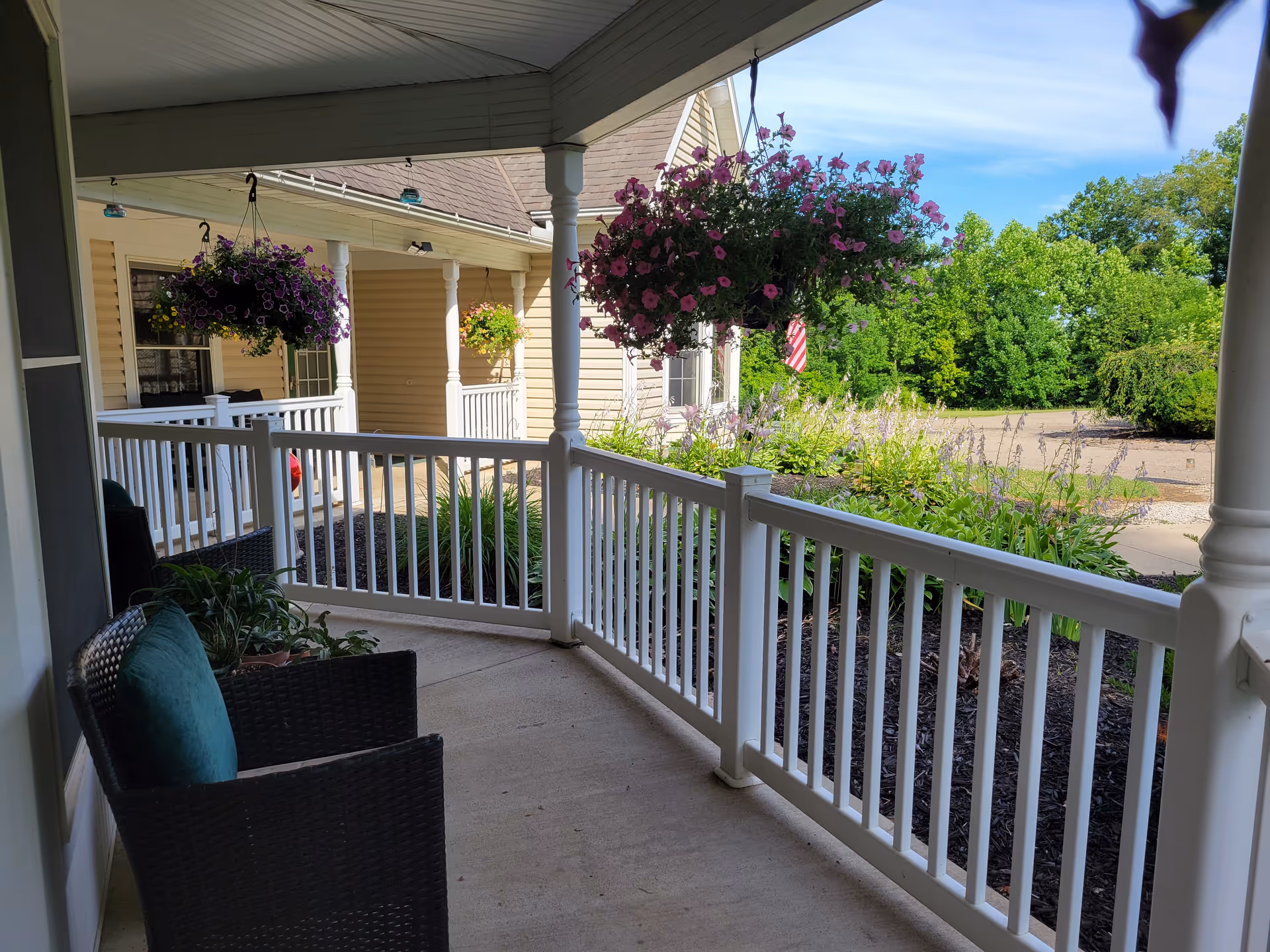 Covered porch area with white railing and two black wicker chairs with green cushions. Hanging flower baskets with purple flowers are suspended from the porch ceiling. The porch overlooks a garden with green plants and a paved driveway. The sky is clear and blue.