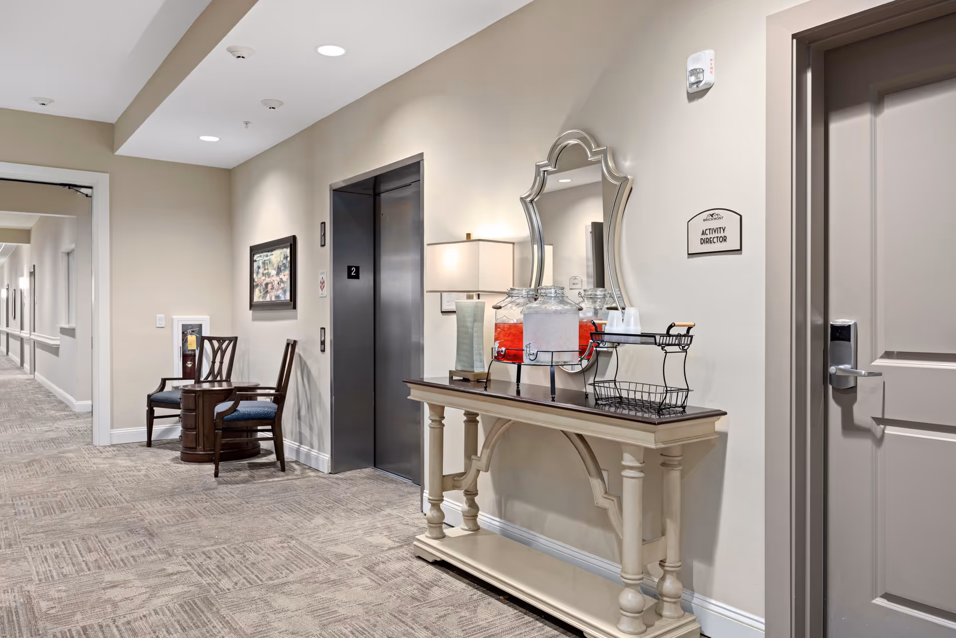 A clean and well-lit hallway in a senior living facility with beige walls and carpeted floor. There is an elevator with the number 2 on it, two wooden chairs with blue cushions next to a small round table, and a decorative console table with a lamp, two beverage dispensers, and a mirror above it. A sign on the wall reads 'Activity Director'.