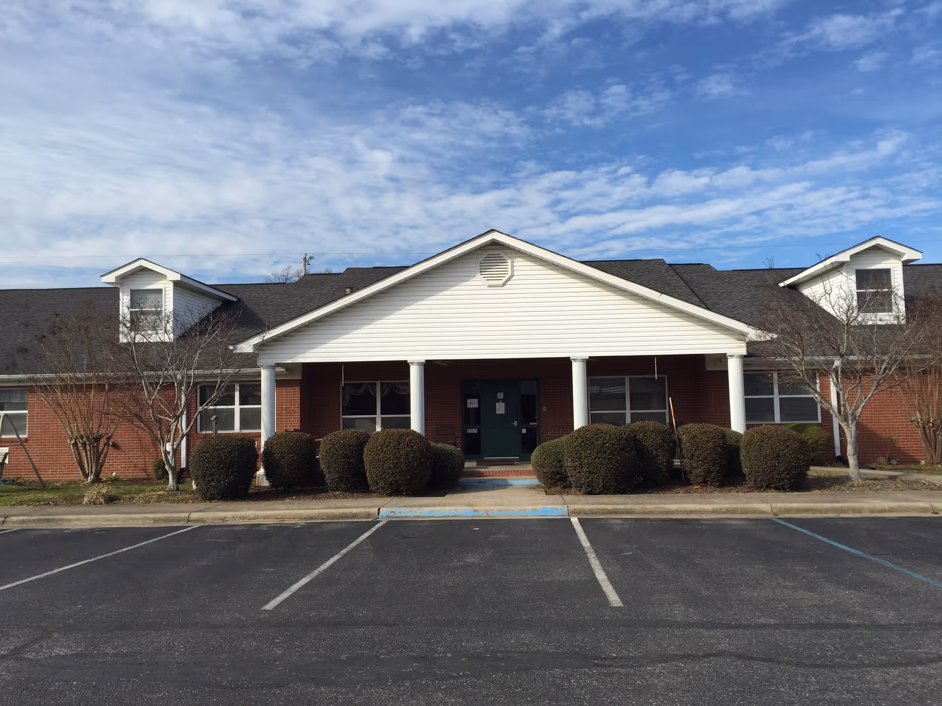 Front exterior view of a single-story brick building with white columns and a peaked roof. The entrance is centered with bushes lining the front and a parking lot in the foreground under a partly cloudy sky.