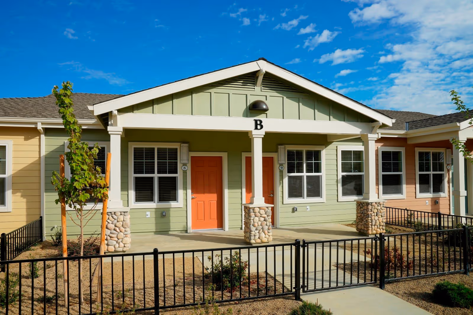 Exterior view of a single-story senior living facility building with green and beige siding, two orange front doors labeled 8 and 9, a small porch with stone pillars, and a black metal fence surrounding the front yard under a blue sky with scattered clouds.