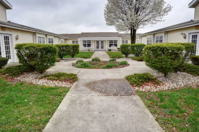 Concrete walkway through a landscaped courtyard leading to a single-story white-siding building with French doors.