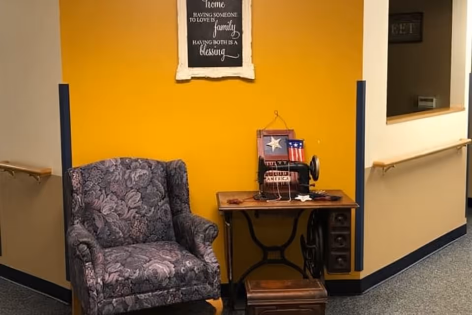 A cozy corner in a senior living facility with a patterned armchair next to a vintage sewing machine on a wooden table. The wall behind is painted yellow and has a framed sign with an inspirational quote about family and love.
